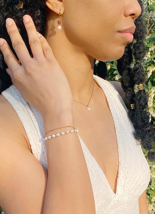 A close-up lifestyle shot of a woman wearing a delicate double-strand pearl bracelet on her wrist, a thin gold necklace with a small white stone, and a gold drop earring with a white pearl, set against a background of green foliage.